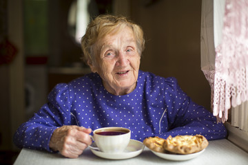 Grandmother, elderly woman, drinking tea sitting at the table in the house.