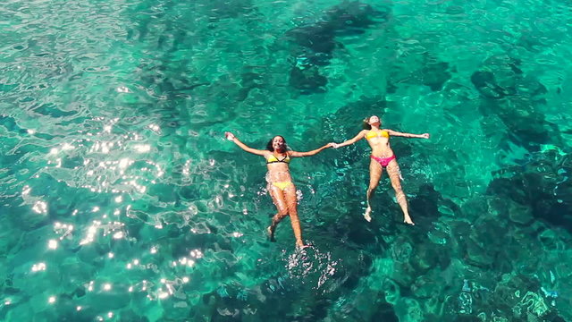 Aerial View of Beautiful young women floating swimming in bikinis holding hands, lying on a water surface in crystal clear ocean. Aerial Perspective. Summer Fun Lifestyle.