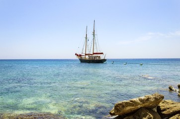A traditional wooden sail boat anchored on the coast of Mykonos,Greece.Wind sails are down and it is tied on rocks by a rope.A very typical greek island summer holiday scene on crystal clear blue sea.
