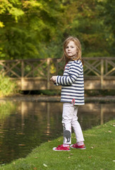 Fototapeta premium Young Girl Looking to Feed the Ducks. A young caucasian girl with ginger hair, is looking to feed the ducks on an ornamental pond in a park. There are vague signs of autumnal colour in the background.