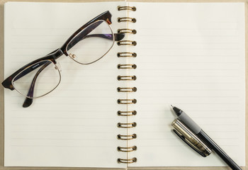 Black pen and notebook with eyeglasses on wood background under