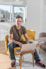 Portrait of a middle aged grey hair man with beard in a stylish vintage living room with wooden floor. He is sitting in an orange chair with his laptop in front of him He is wearing casual clothes