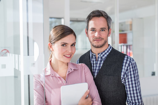 Portrait Of Two Smiling Coworkers Looking At Camera. A Woman With Pink Shirt Holding A Tablet And A Grey Hair Man With Beard And A Blue Checkered Shirt, Are Standing In Front Of A Glass Door.