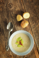 Top view of a potato soup in a blue bowl with fresh potatoes and spoon on a wooden background