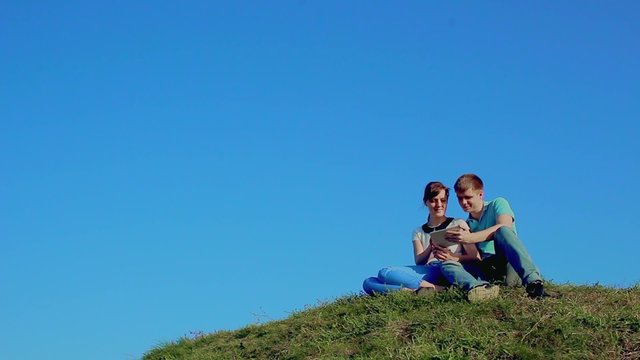 Young loving couple sitting on grass on the hill and using tablet pc computer. Clear blue sky background 