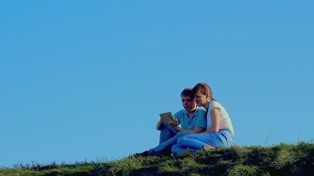 Young loving couple sitting on grass on the hill and using tablet pc computer. Clear blue sky background 