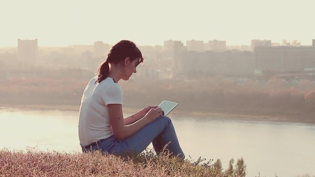 Young woman sitting on the hill with tablet pc in hands. Evening cityscape background 