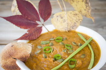 horizontal close up image of a bowl of pumpkin soup with green onion and pumpkin seeds on top and some fall leaves in the background.