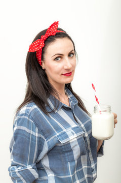 Mature brunette Caucasian woman wearing a blue flannel shirt and a red bow on her head is drinking a glass of milk with a white and red straw from a clear glass.