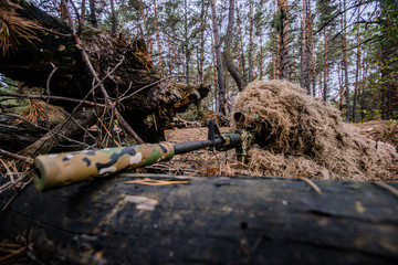 Camouflaged sniper lying in forest and aiming through his scope/Sniper in camouflage suit is hiding and aiming behind a tree