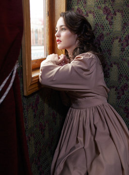 Young woman in beige vintage dress looking trough the  window in