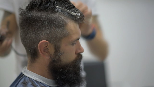 stylish hairdresser with tattoos on his arms makes cut and style hair brutal bearded man in the barbershop