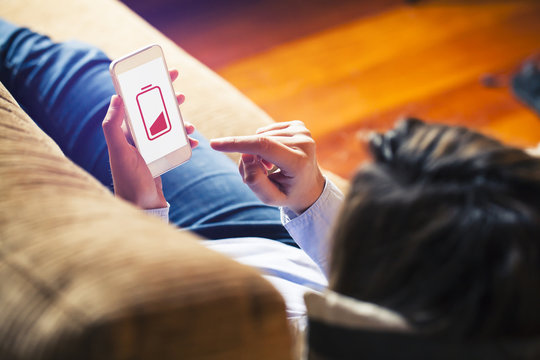 Low battery. Woman lying on a sofa, touching mobile screen with red low battery icon.