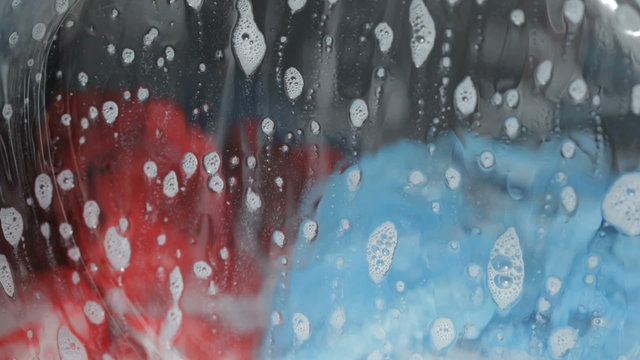 Closeup of a front-loading washing machine filled with brightly colored tumbling clothing. Shallow focus on soap bubbles.