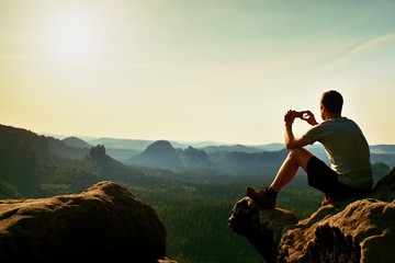 Un touriste en t-shirt gris prend des photos avec un téléphone intelligent au sommet du rocher. Paysage vallonné de rêve ci-dessous, lever de soleil brumeux rose orange dans une belle vallée au-dessous des montagnes rocheuses.
