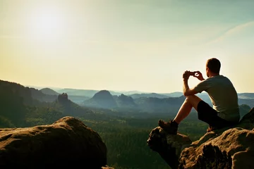 Tuinposter Toerist in grijs t-shirt maakt foto& 39 s met smartphone op de top van de rots. Dromerig heuvelachtig landschap hieronder, oranje roze mistige zonsopgang in een prachtige vallei onder rotsachtige bergen. © rdonar