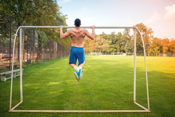 Young muscular man doing pull ups on football court, hardcore training outdoors