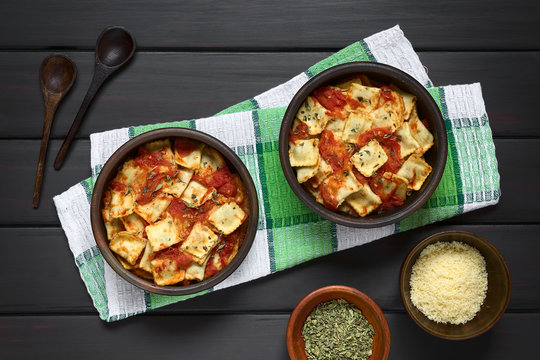 Baked ravioli with homemade tomato sauce in rustic bowls with grated cheese and dried oregano in small bowls, wooden spoons on the side, photographed overhead on dark wood with natural light