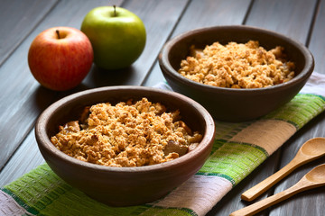 Baked apple crumble or crisp on kitchen towel, wooden spoons and fresh apples on the side, photographed on dark wood with natural light (Selective Focus, Focus one third into the first crumble)