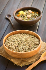 Raw lentils (lat. Lens culinaris) in wooden bowl with lentil soup in the back, photographed on dark wood with natural light (Selective Focus, Focus one third into raw lentils)