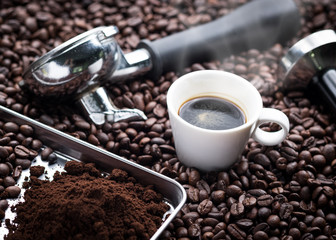 A cup of hot and fresh espresso coffee lay on many roast coffee beans also see an espresso machine group head, coffee tamper and ground coffee lay on many roasted coffee beans.