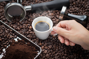 Fresh espresso. Hand grabbing an ear cup of of hot espresso coffee also see an espresso machine group head, coffee tamper and ground coffee lay on many roasted coffee beans.