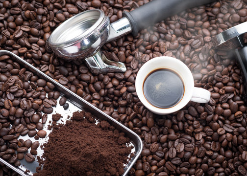 A cup of hot and fresh espresso coffee lay on many roast coffee beans also see an espresso machine group head, coffee tamper and ground coffee lay on many roasted coffee beans.