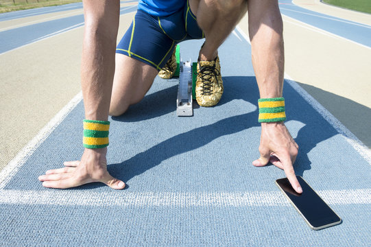 Athlete crouching at the starting line of a running track wearing Brazil colors wristbands checking his mobile phone
