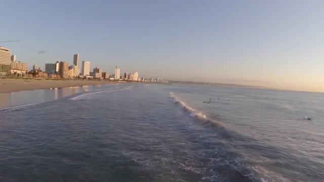 Aerial over Durban's surf with waves breaking, surfers in the water and city skyline on the horizon at sunrise.