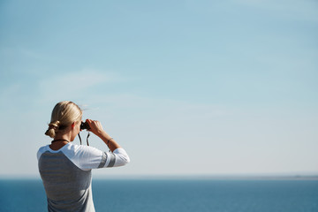 Woman tourist looking through binoculars at distant sea, enjoying landscape, copy space
