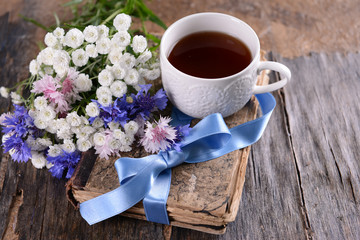Old book with beautiful flowers and cup of tea on wooden table close up
