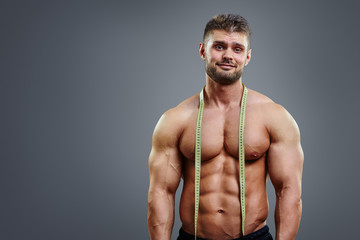 Shy bodybuilder with tape measure standing isolated over gray background. Studio portrait of cute...