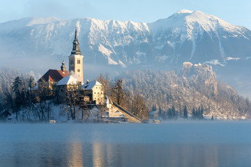 Church on lake Bled with boats arriving to the stairs and castle and snowy mountains in the background