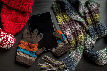 the tablet is on the gloves, as if lying in the hands of the person near to a black wooden table are other colorful winter clothes: a red hat and scarf