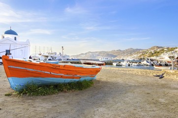 Colourful fishing and sail boats anchored on the Chora port in Mykonos,Greece. View of orange,blue boat on sand shore and the whitewashed blue dome church of Hora harbour, a greek island landscape. © f8grapher