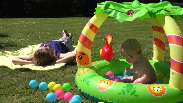Careless mother woman lay on plaid and baby splash water in colorful kiddie pool on garden meadow. Static shot. 4K 
