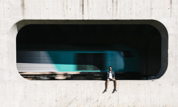 Spain, Tarragona, young man sitting in niche of a concrete wall with moving train in background