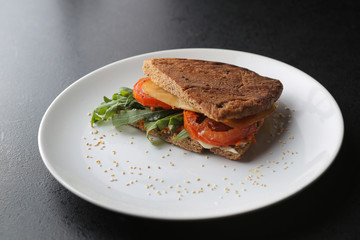 Vegetarian sandwich with tomato, arugula and cheese on white plate on dark background.