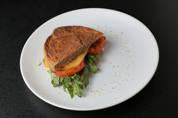 Vegetarian sandwich with tomato, arugula and cheese on white plate on dark background.