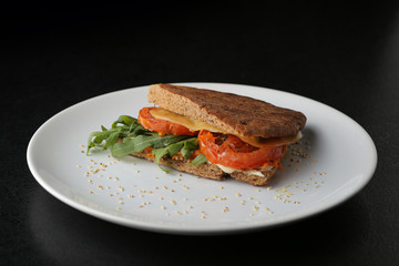 Vegetarian sandwich with tomato, arugula and cheese on white plate on dark background.