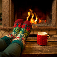 Feet in woollen socks by the Christmas fireplace. Woman relaxes by warm fire with a cup of hot...