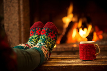 Feet in woollen socks by the Christmas fireplace. Woman relaxes by warm fire with a cup of hot drink and warming up her feet in woollen socks. Close up on feet. Winter and Christmas holidays concept. - Powered by Adobe