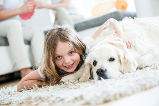 Little girl cuddling with her dog, lying on floor, parents in background