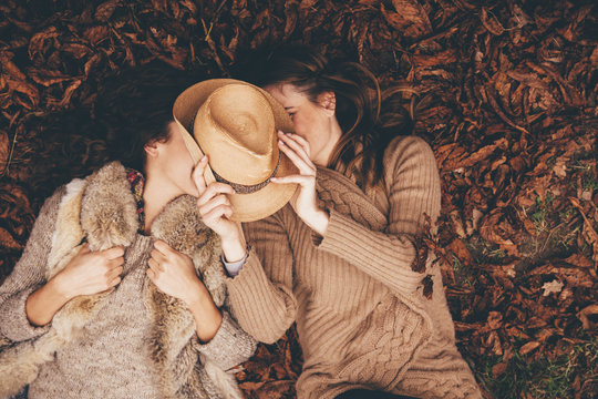 Two female friends lying side by side on autumn leaves in an park hiding behind a hat