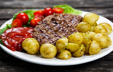 Fresh juicy grill burgers, served with ketchup, young potatoes, tomatoes and vegetables. On wooden table