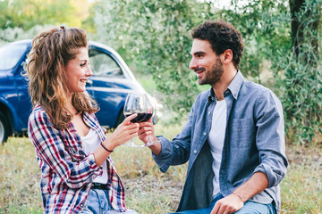A couple of young lovers drinking red wine from a glass goblet at a picnic in the countryside in Tuscany, Italy. Sitting on lawn among the olive trees on a late summer day, behind them an old blue car