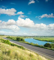asphalt road and river in green landscape under clouds in blue s