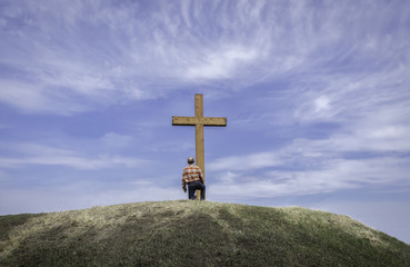 horizontal image of a man kneeling by a wooden cross on a grassy hill surrounded by a beautiful...