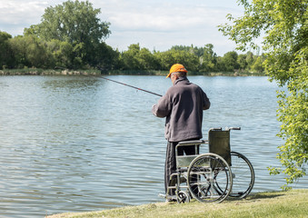 image of a homeless man standing next to his wheel chair by the lake with a fishing rod in the water on a warm summer morning,