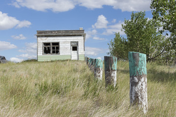 horizontal image of an old abandoned business building with fence posts sitting amidst tall grass that has not been mowed in the summer time.
